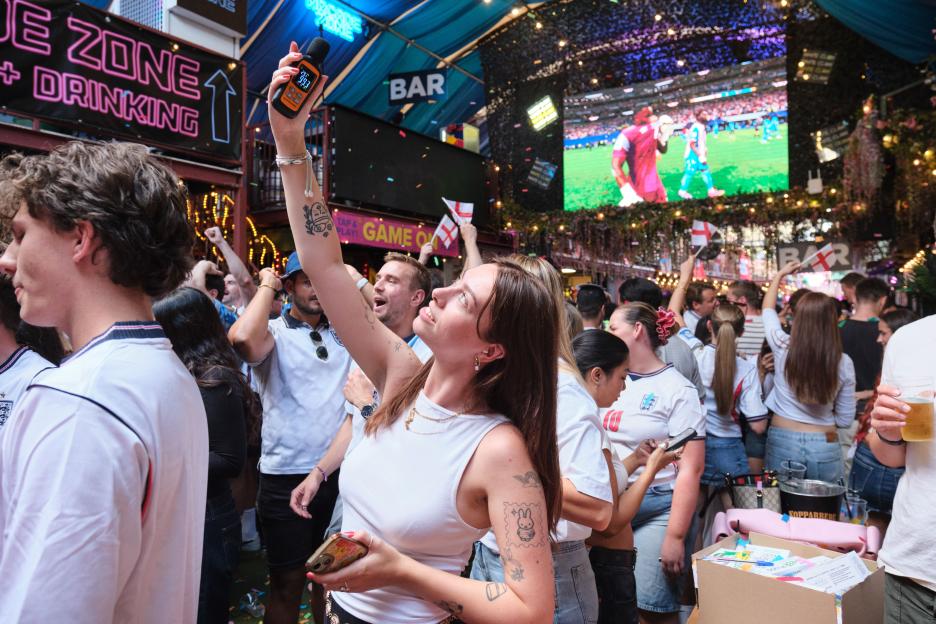 A woman holds up a sound level meter measuring 99.3 decibels while watching a soccer game on a large screen with a crowd.