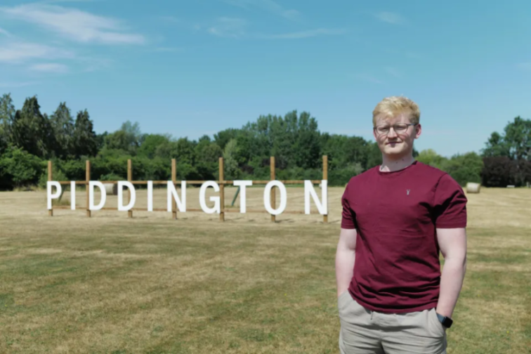 A young man in a red shirt and glasses stands in a field in front of a "PIDDINGTON" sign.