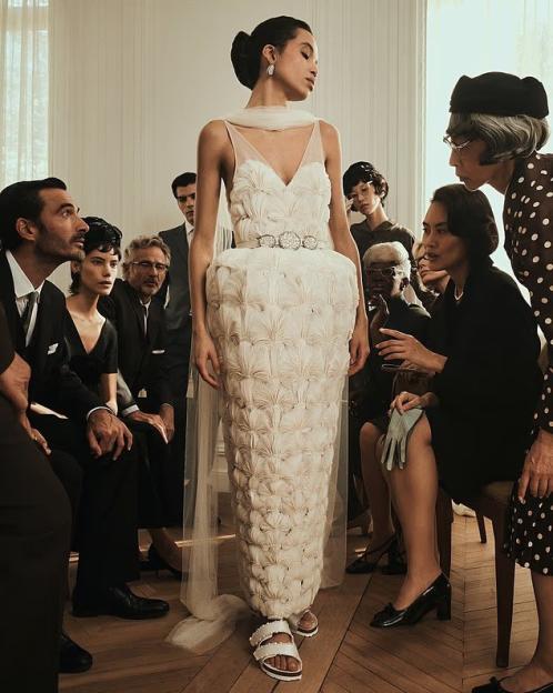 A model in a white bridal gown with a voluminous skirt and white Birkenstock sandals stands surrounded by seated onlookers.