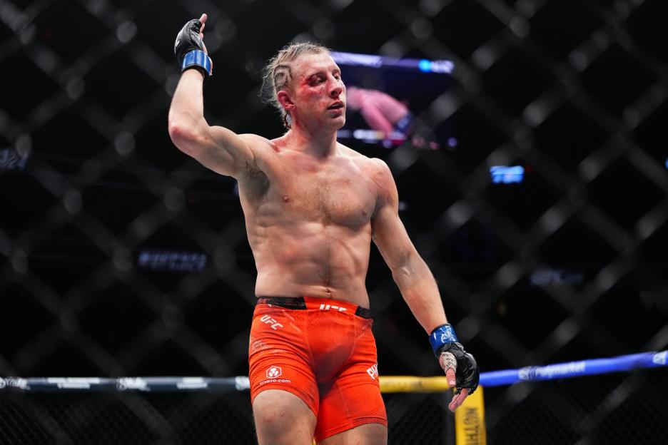 Justin Gaethje with a bloodied face, wearing red shorts and black MMA gloves, raises his arm in the UFC octagon.