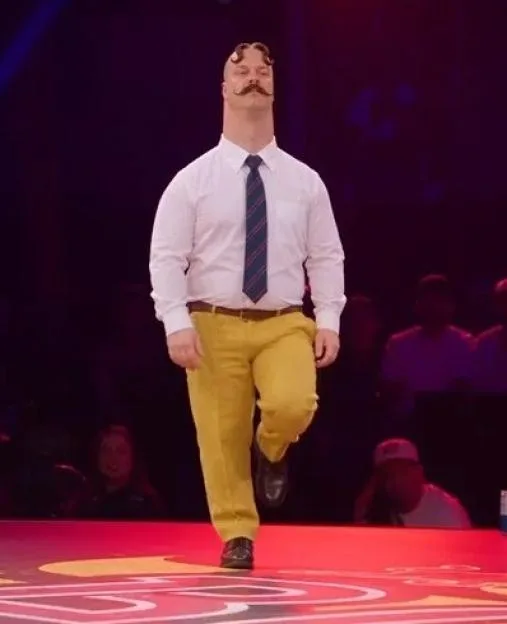 A man with a large mustache and styled hair walks on a stage wearing a white shirt, striped tie, and yellow trousers.
