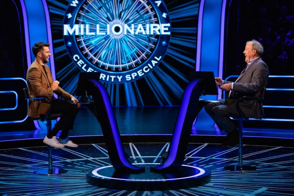 Two men sit on high chairs opposite each other on the "Who Wants to Be a Millionaire Celebrity Special" set.