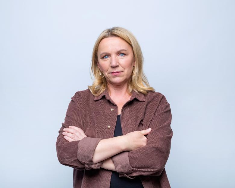 Portrait of a worried mature woman with blonde hair and blue eyes, wearing a brown shirt and standing with arms crossed.