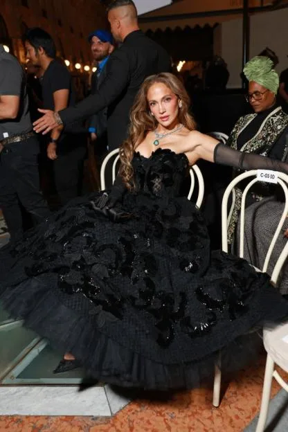 Jennifer Lopez sitting in a black strapless gown with a full skirt adorned with lace and sequins, holding a black clutch.