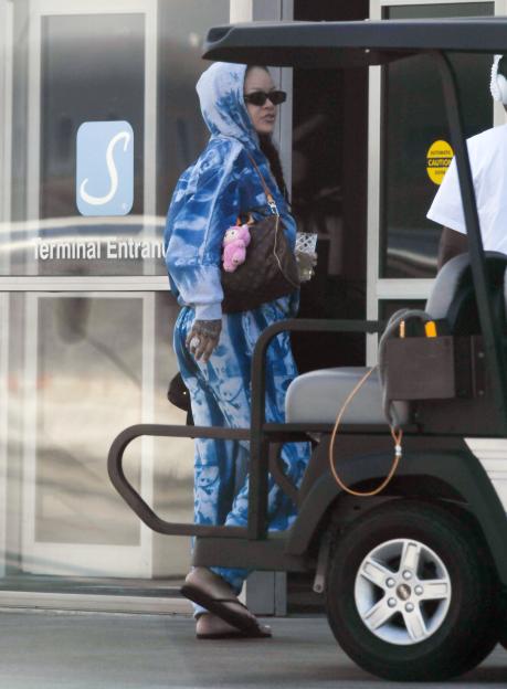 Rihanna in a blue sweatsuit carrying a Louis Vuitton bag and a clear cup, walking past a golf cart at an airport.