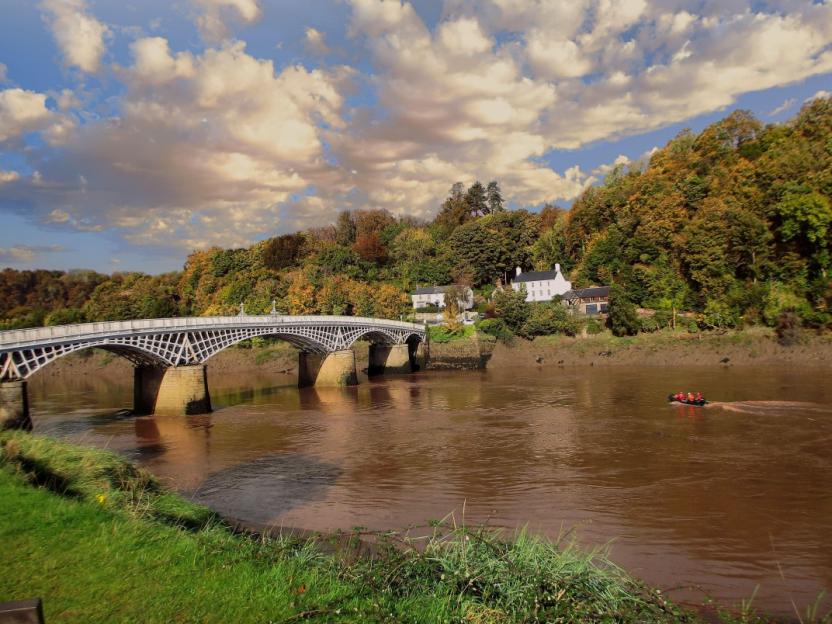 White-painted bridge with intricate ironwork spanning a brown river, with a small boat, houses, and trees on the far bank under a cloudy sky.