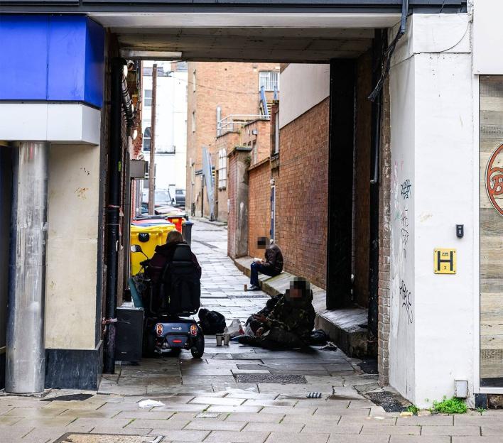 Two people sitting in an alleyway, one in a motorized wheelchair and another on the ground.