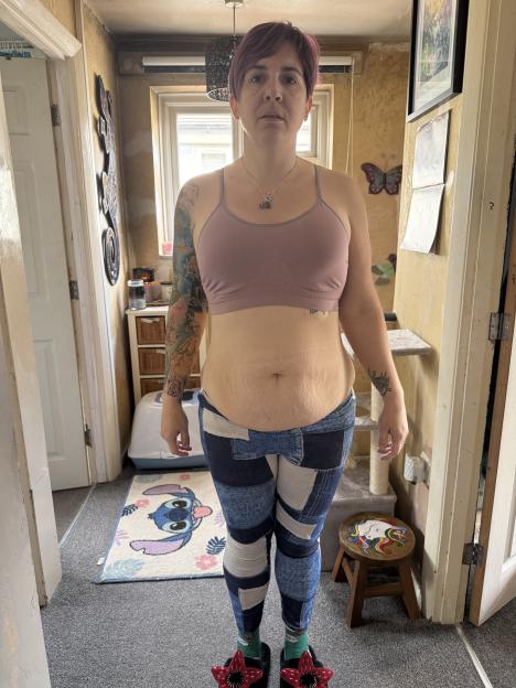 Tammy Downing, 44, stands in a room wearing a sports bra, patterned leggings, and slippers.