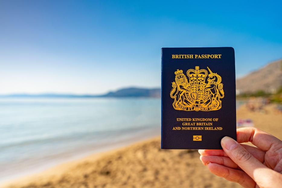A hand holding a British passport against a bright blue sky and beach background.