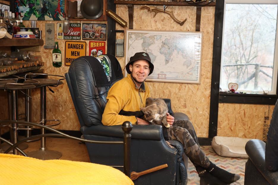 Sam Griffiss sits in a black chair with a cat on his lap inside his home, which is made of wooden panels and decorated with signs and maps.