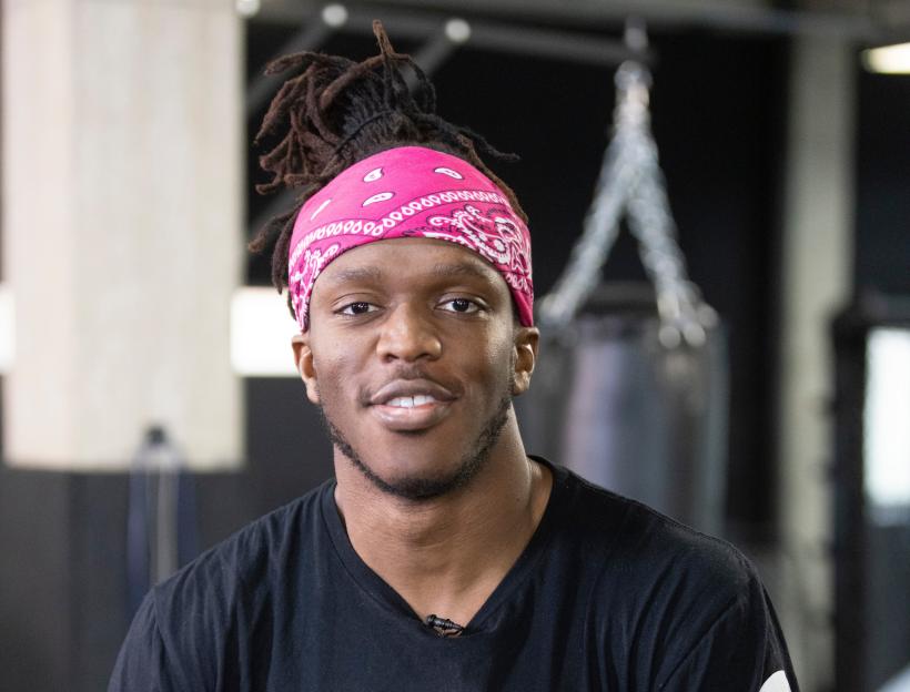 KSI (Olajide Olatunji) in a black "London Shootfighters" t-shirt and a pink bandana, leaning on boxing ropes.