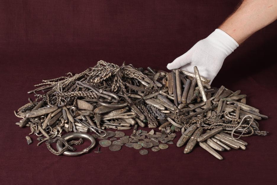 Gloved hand placing silver Viking hoard of bracelets, coins, and bars on a red cloth.