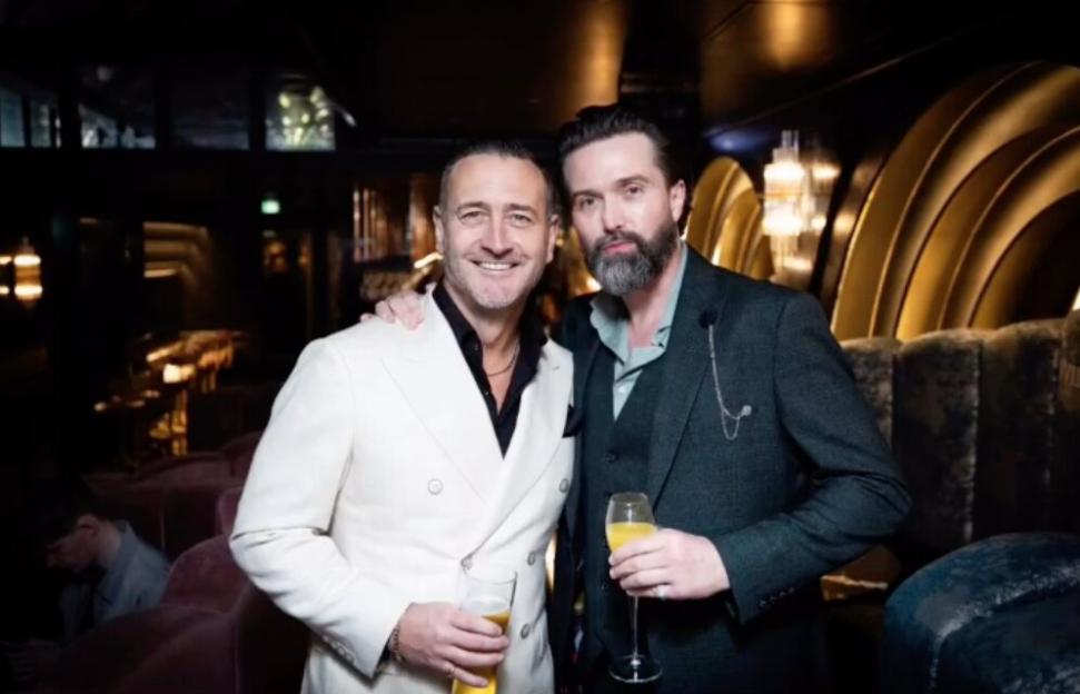 Will Mellor and a Hollyoaks co-star pose together with drinks in a dimly lit venue.