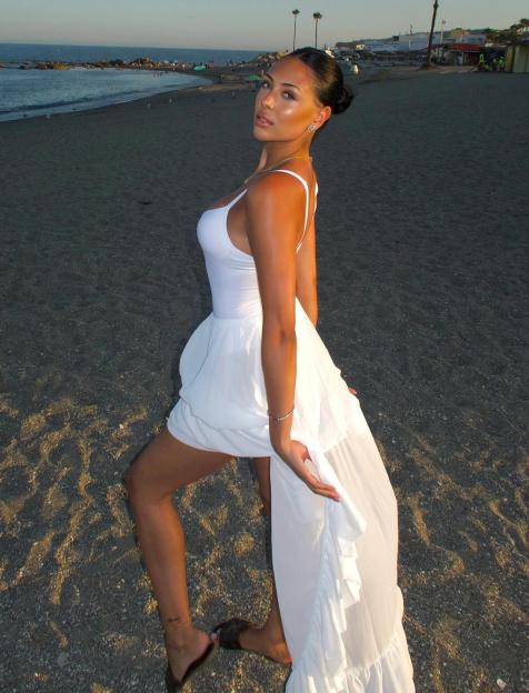 A young woman in a white dress stands on a sandy beach looking back at the camera.