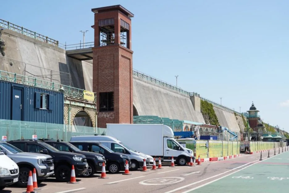 A street-level view shows a row of parked cars and vans alongside a road with a green cycle lane, next to a tall brick lift tower and a concrete seawall under construction.