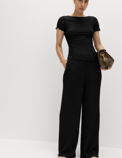 A woman wearing a black short-sleeved top, black wide-legged pants, and holding a brown purse.