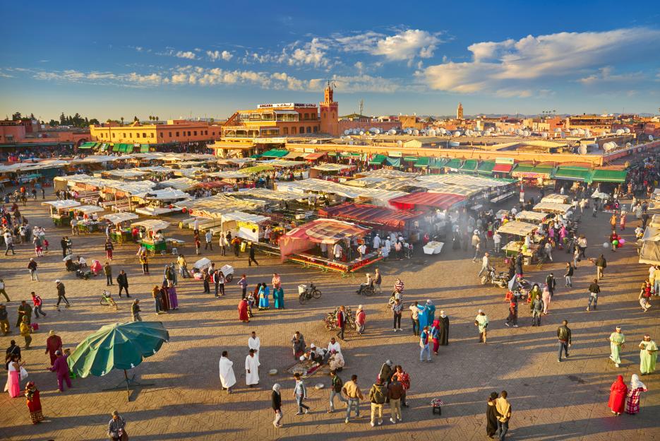 Djemaa el-Fna Square in Marrakech Medina, Morocco, bustling with people and market stalls under a bright sky.