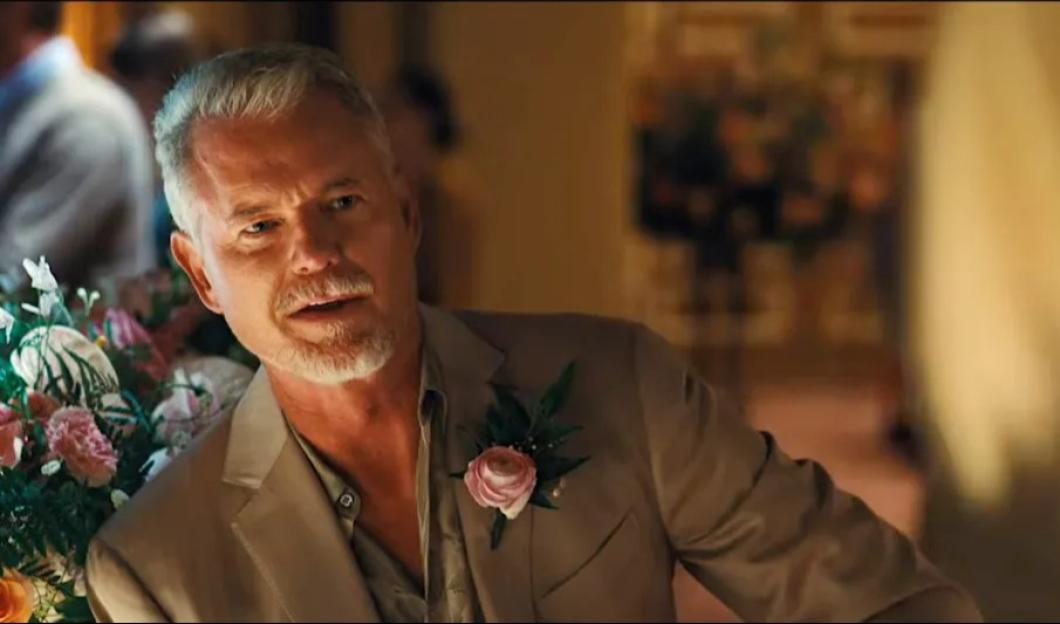 A man with gray hair and a beard, wearing a tan suit with a pink flower boutonnière, looks forward.
