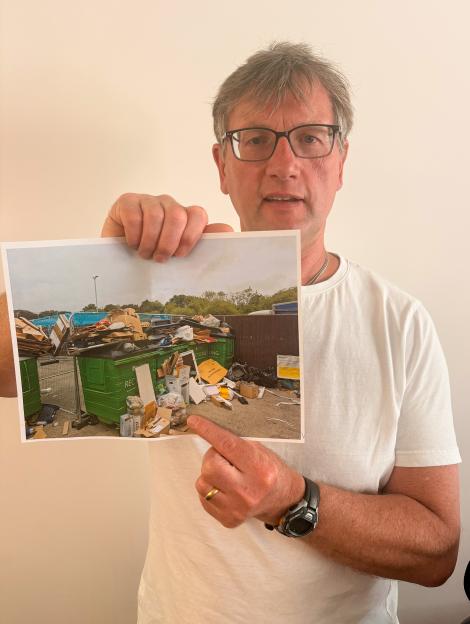 A man wearing glasses and a white t-shirt holds up a printed photo of an overflowing recycling facility.
