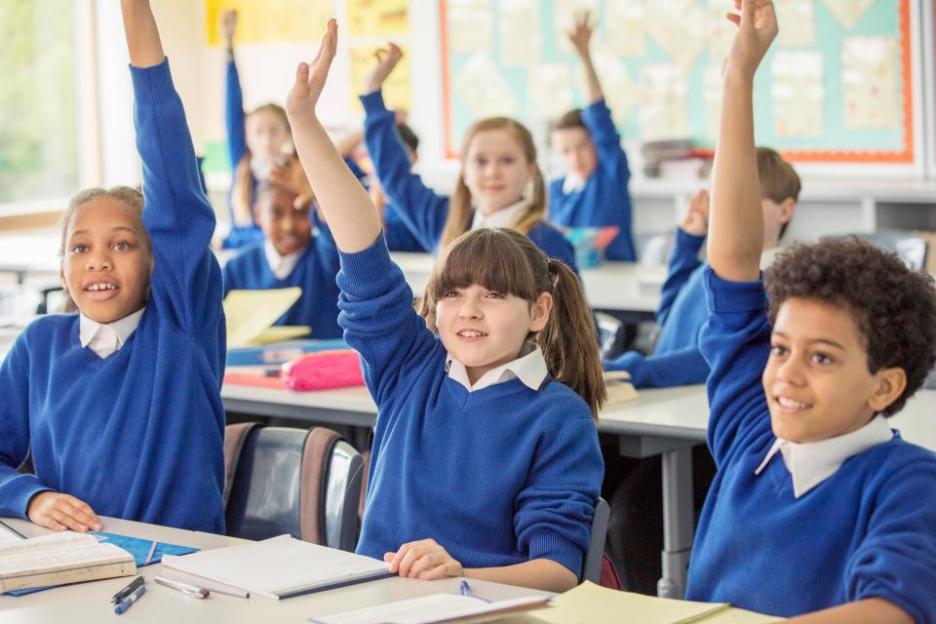Children in blue school uniforms raise their hands in a classroom.