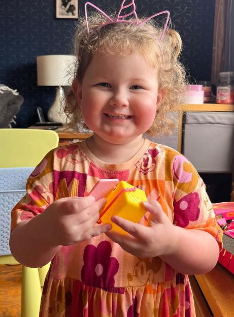 A young girl with curly blonde hair and rosy cheeks wearing a pink unicorn headband and a colorful dress, holding a yellow toy, smiles at the camera.