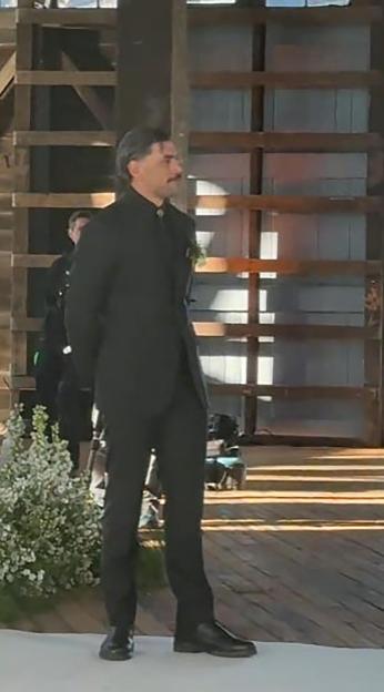 A man in a black suit with a mustache stands alone on a set, waiting.