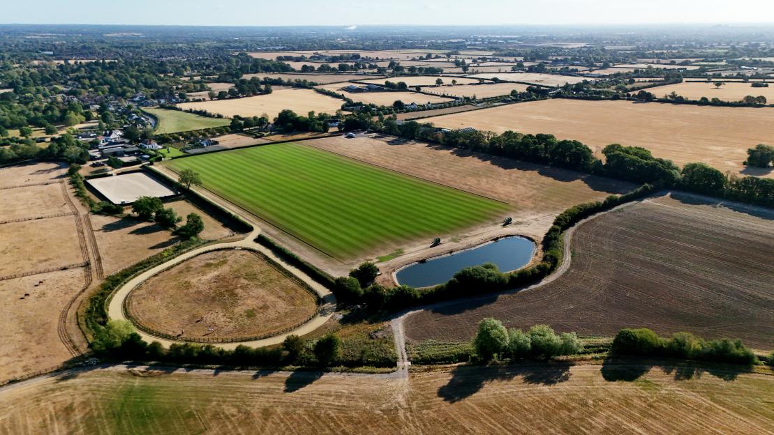 Aerial view of the King Power Polo pitch in Holyport, Berkshire, showing a vibrant green polo field, surrounding dry, brown fields, a small pond, and distant buildings, highlighting drought conditions.