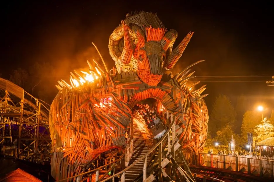 A roller coaster train exiting a giant wooden monster structure with ram horns illuminated by orange lights at night.