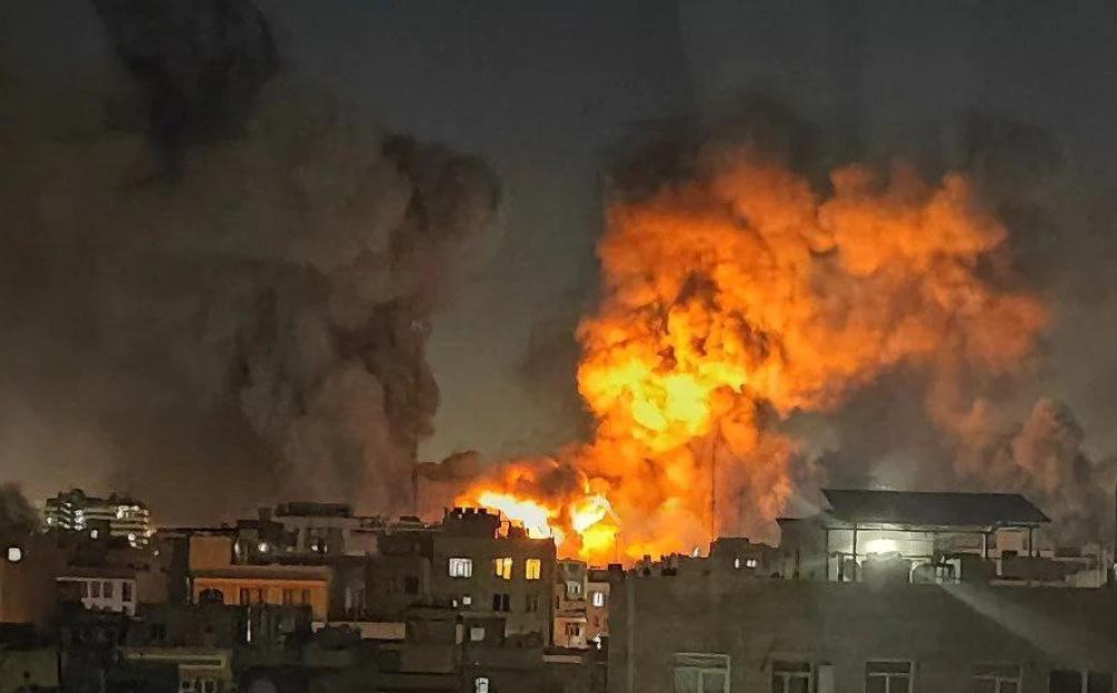 Large explosion with orange flames and dark smoke billowing into the night sky above a cityscape.