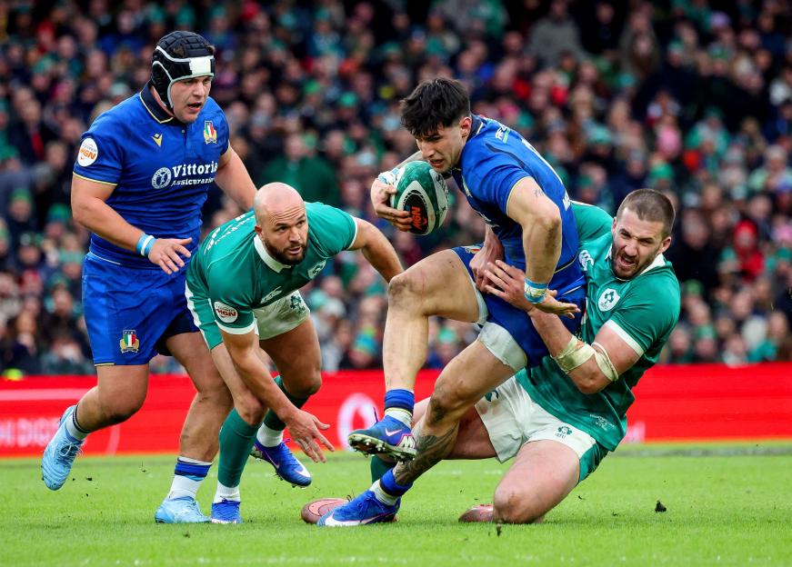 An Italian rugby player in a blue jersey, with the ball in his hands, is tackled by two Irish players in green jerseys.