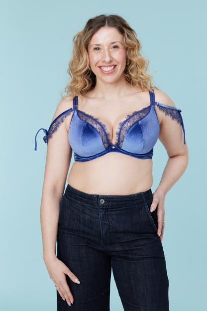 A smiling woman in a blue velvet bra with lace trim and dark jeans poses against a blue background.