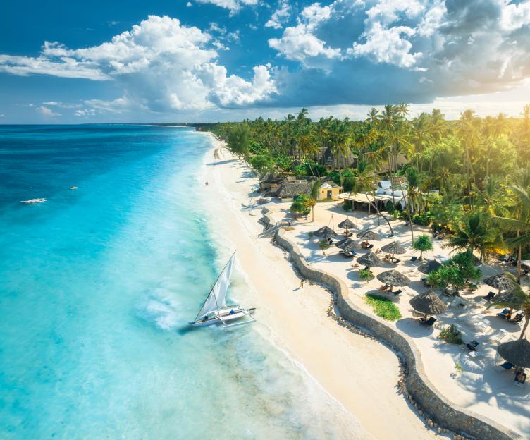 Aerial view of a sailboat on the blue sea near an empty white sandy beach with umbrellas and palm trees at sunset in Zanzibar.
