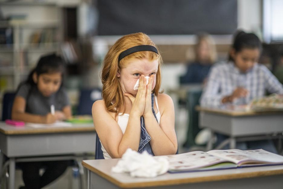 A red-haired girl in a classroom sneezes into a tissue.