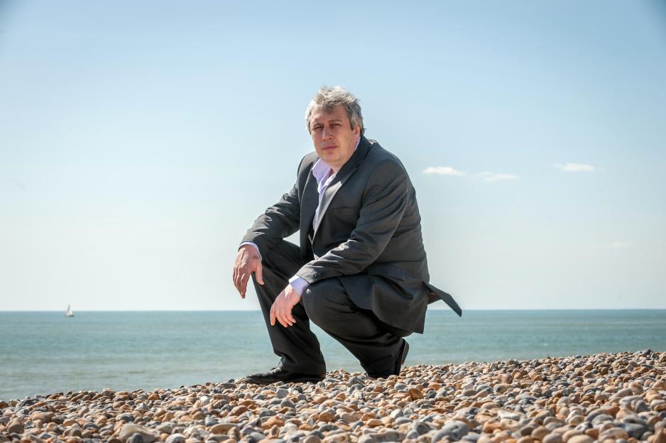 Newspaper columnist Rod Liddle in a suit, squatting on a rocky beach.