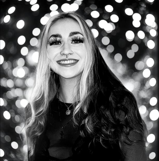 A black and white portrait of a smiling woman with long hair, piercings, and dramatic eyelashes, with blurred lights in the background.