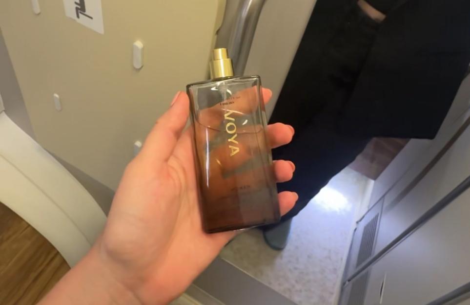 A hand holding a bottle of Moya perfume inside an airplane lavatory.