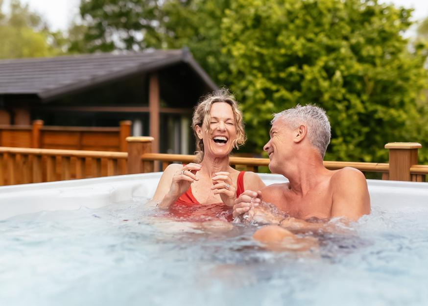 A laughing couple enjoying a hot tub on a wooden deck with a cabin and trees in the background.