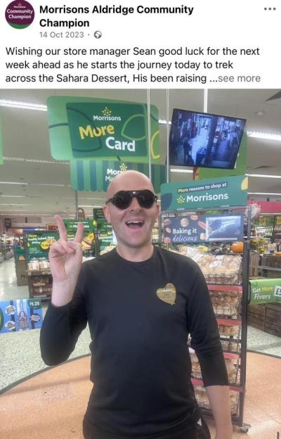 Sean, the bald store manager, wearing black sunglasses, smiles and makes a peace sign in a Morrisons store, ready to trek the Sahara Desert.