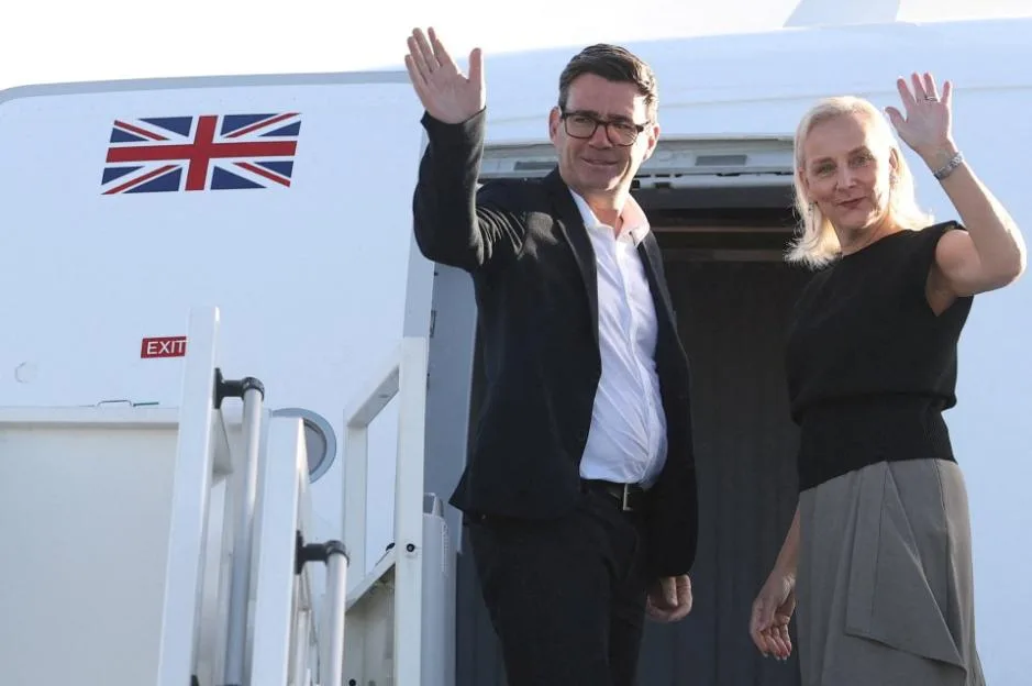 British Prime Minister Andy Burnham and his wife Marie-France van Heel wave from the steps of a plane.