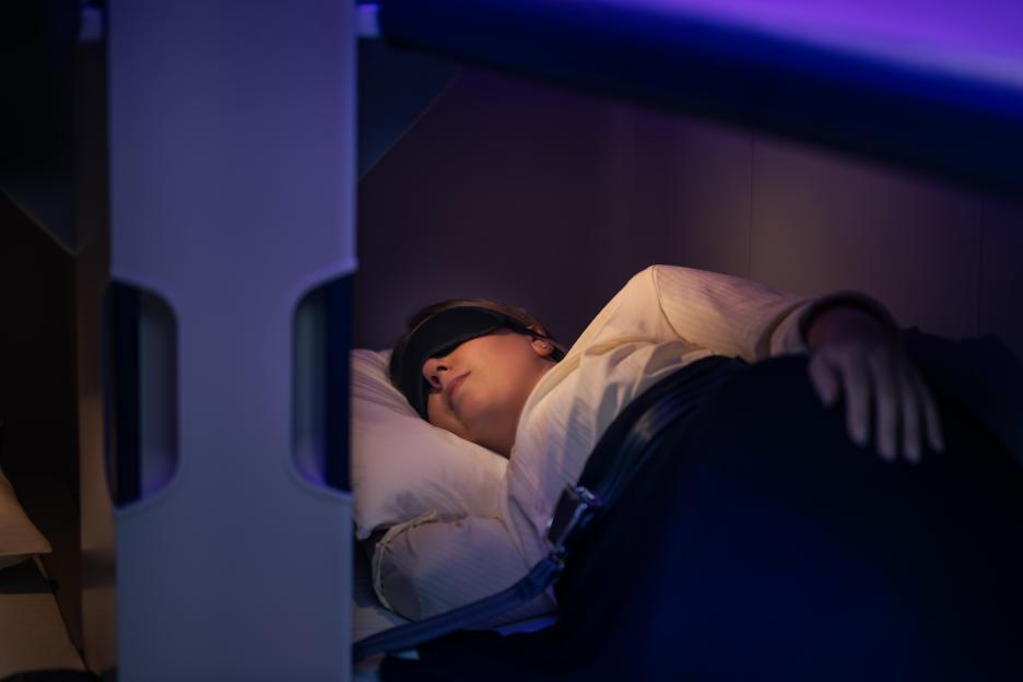 A person wearing an eye mask sleeps in an airline bunk bed.