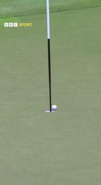 A golf ball rests on the green next to a hole, with the flagstick extending into the hole.