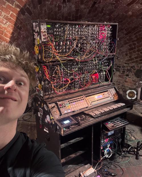A man taking a selfie next to a large modular synthesizer setup.