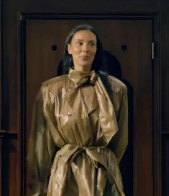 Maya Jama wearing a shiny gold trench coat in front of a dark wooden door.