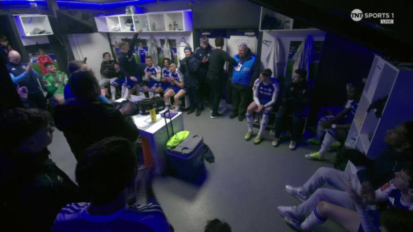 Football players and staff in a locker room, clapping for someone off-camera.