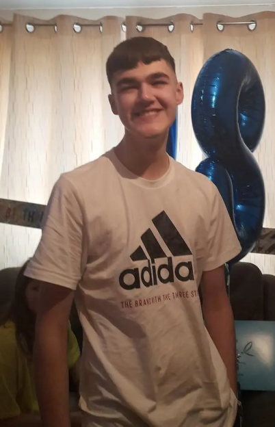 A young man wearing a white Adidas t-shirt, smiling, with blue balloons in the background.