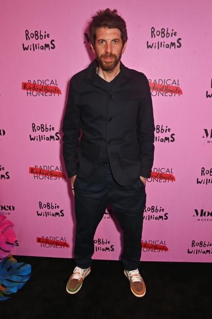 A man in a dark jacket and trousers stands against a pink wall with Robbie Williams' "Radical Honesty" exhibition branding.
