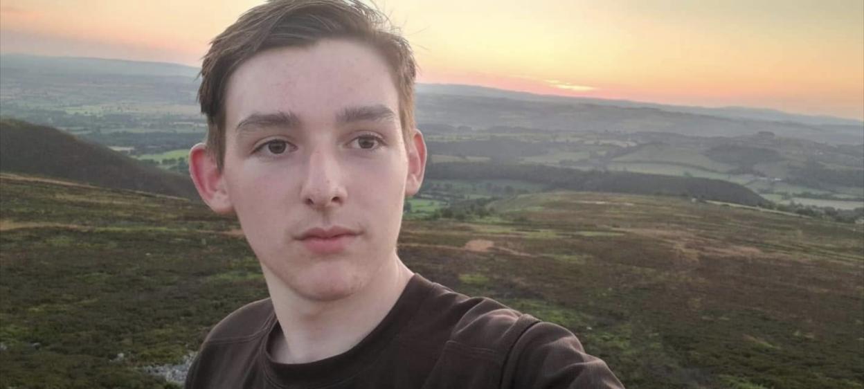 A young man, Benjamin Worrall, stands on a hillside with a valley and sunset behind him.