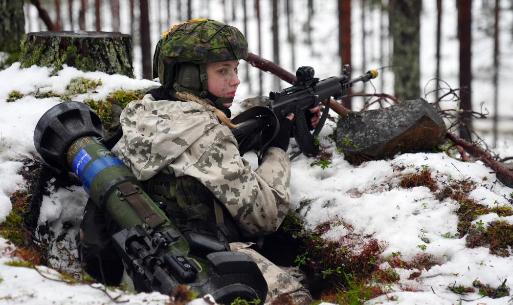Finnish soldier in camouflage uniform, helmet, and ear protection, holding a rifle and lying in a snowy trench next to a Javelin anti-tank missile system.
