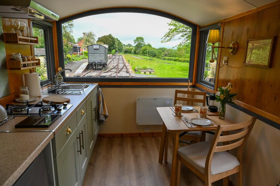 The interior of a renovated train carriage at Rowden Mill Station, featuring a small kitchen and dining area with a window overlooking train tracks and a brake van.