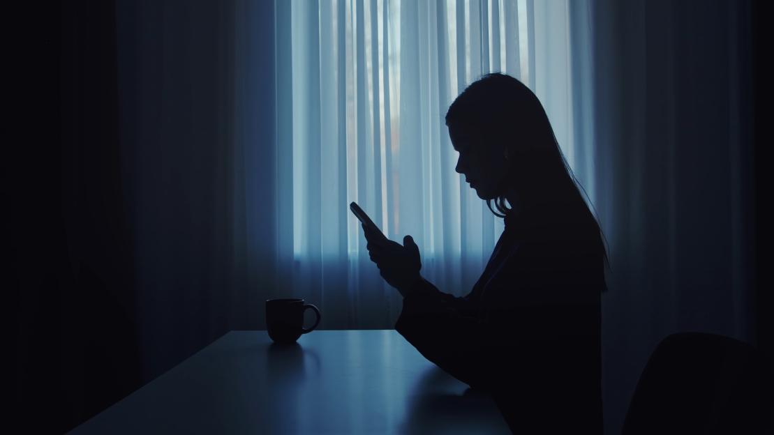 Silhouette of a woman using her smartphone at a desk with a coffee cup in a dimly lit room.